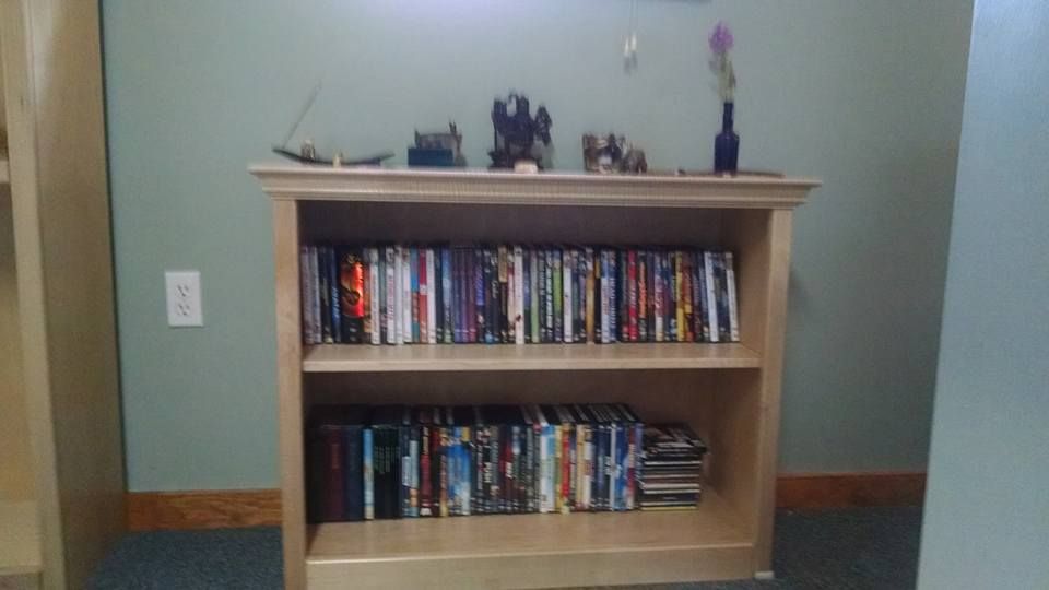 Maple bookshelf with two shelves loaded with dvds and a few cds and video games. On top of the shelf resides an alter, including an arrangement of a glass bottle filled with protective herbs and with a small silver key hanging from white ribbon from the neck of the bottle, a stone incense burner, statue of Kali, indigo glass bottle, two raku fired candle holders, a wood box stained blue with a pentacle carved on the lid, a raku fired horse with copper wire mane and tail, a willow wand wrapped in copper wire with an amethyst stones on either end, and a scattering of smaller rocks, pendants and other items.