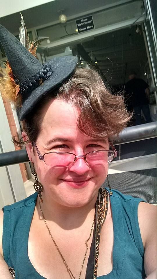 A white person with short wavy dark brown hair wearing a small felt witch's hat decorated with orange feathers and an orange leaf, brass gear earrings and a dark blue tank top smiling while her glasses slip down their nose.