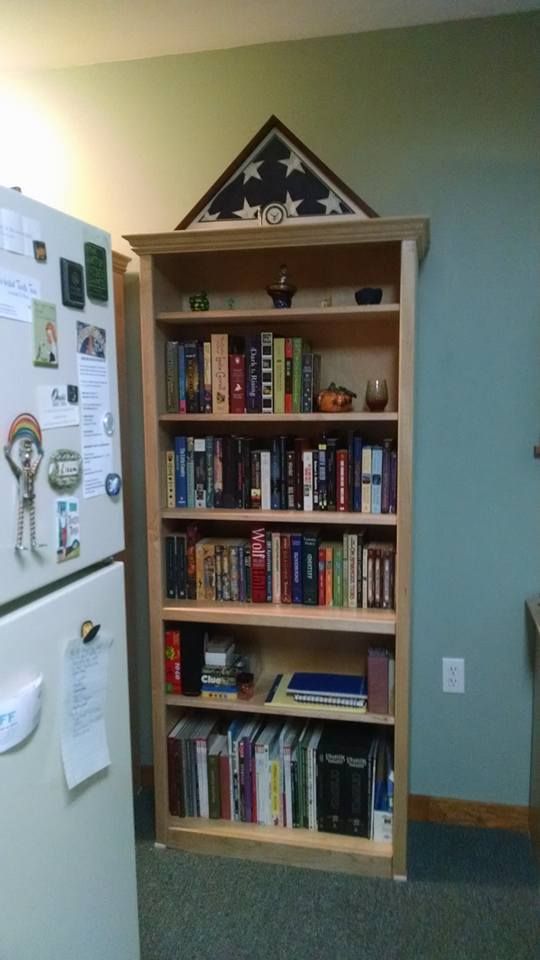 A tall maple bookshelf. On the very top is an American flag in a dark wood frame, with a bullet casing sitting in front of it. The remaining shelves are filled with assorted knick-knacks, books, and games.
